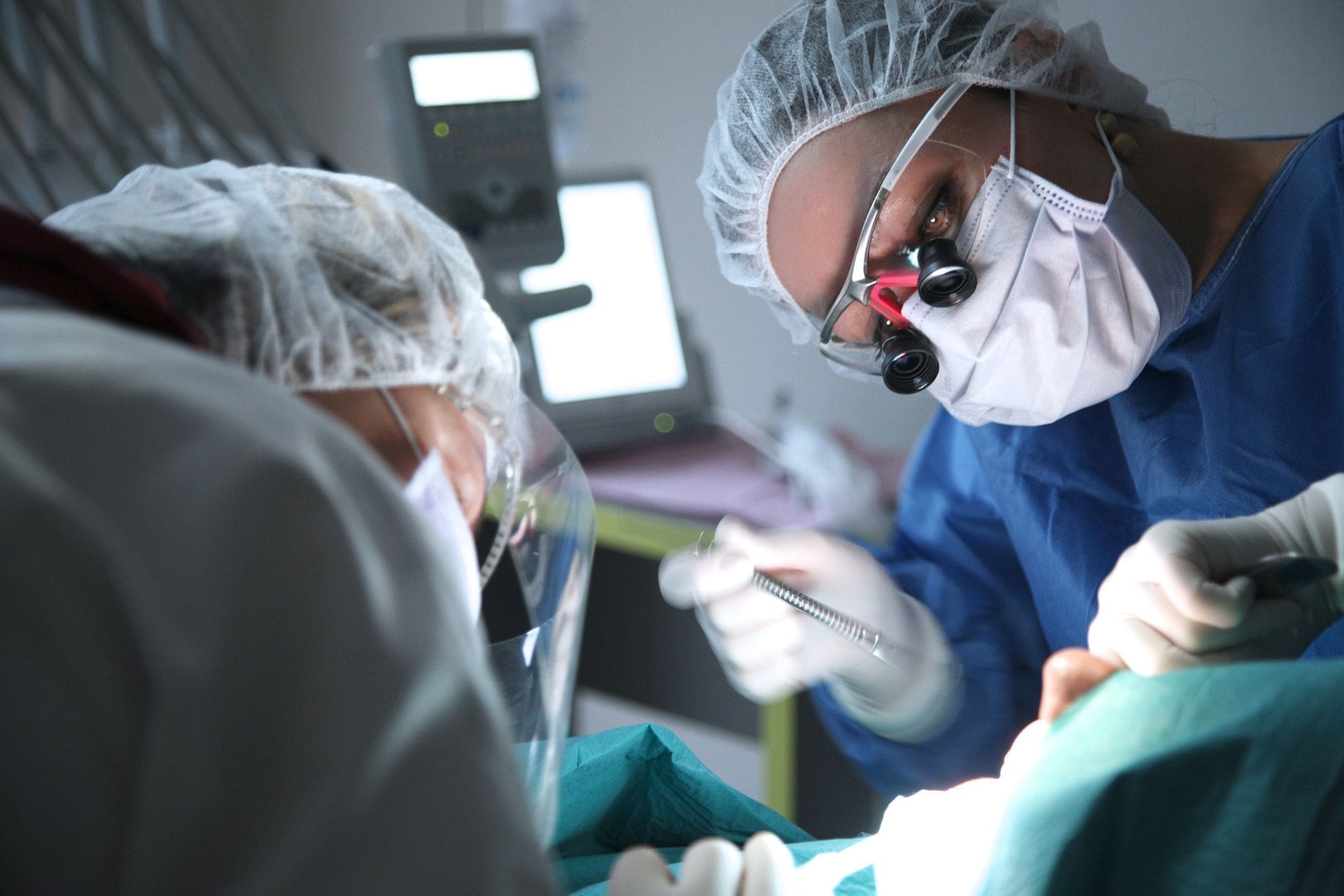 Two medical professionals wearing surgical masks, gowns, and hair covers perform a procedure under bright light, using dental or surgical instruments. One wears magnifying glasses and focuses intently on the patient.