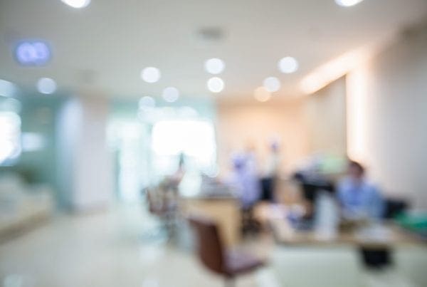 Blurred image of a hospital or clinic reception area with indistinct figures, desks, chairs, and bright lighting, conveying a busy medical or healthcare environment.