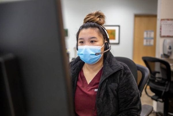 A woman wearing a medical mask and headset sits at a desk, looking at a computer monitor in an office or clinic setting.