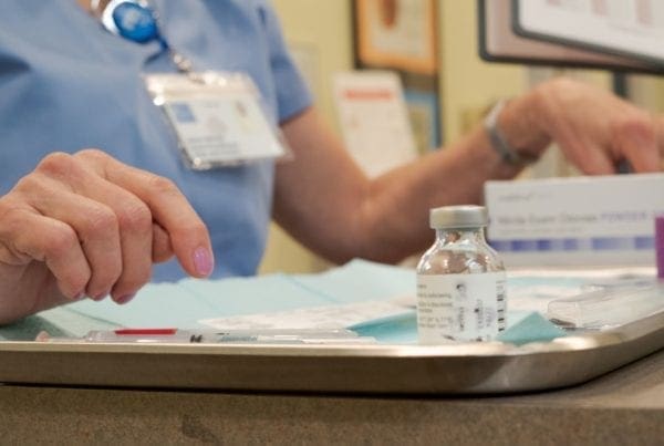 A healthcare worker in scrubs prepares a tray with a vaccine vial, syringes, and alcohol wipes, ready for administration. Only the worker’s hands and torso are visible; their ID badge hangs from their neck.