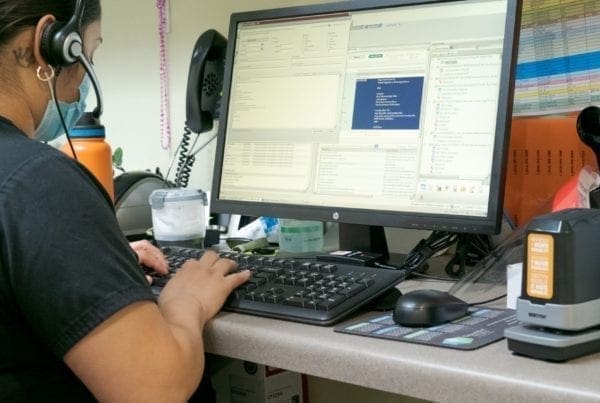 A person wearing a headset works at a desk, typing on a keyboard in front of a large computer monitor displaying various windows and programs. Office supplies and paperwork are visible on the desk.