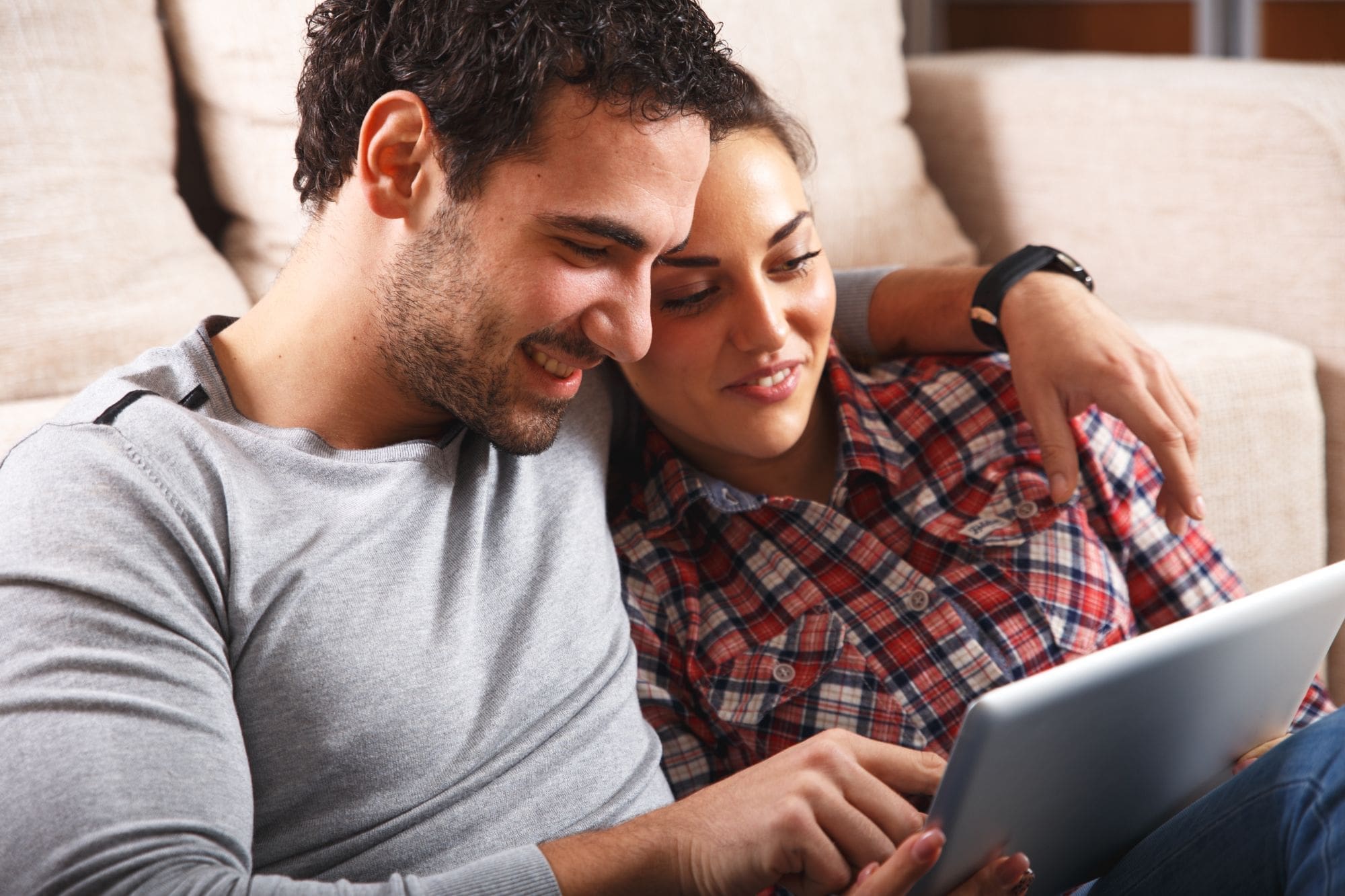 A man and woman sit closely together on a couch, smiling and looking at a tablet screen. The man has his arm around the woman, and both appear relaxed and engaged with the device.