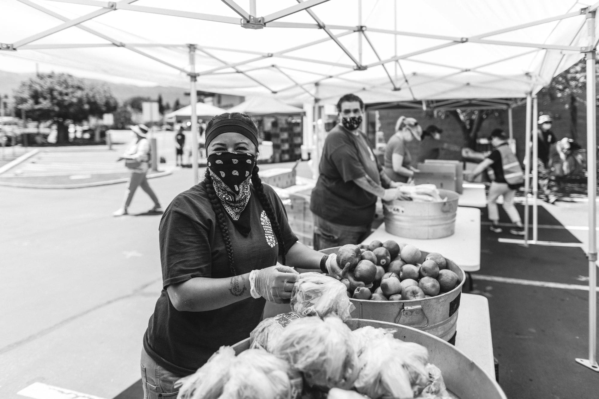 A woman wearing a mask and gloves stands under a canopy at an outdoor food distribution event, smiling as she handles bags of fresh produce. Other volunteers and tables of food are visible in the background.
