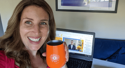A smiling woman holds an orange mug in front of a laptop displaying a man speaking in a video call or webinar. She is indoors, and a framed picture hangs on the wall behind her.