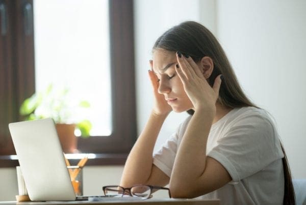 A young woman sits at a desk with a laptop, eyes closed and hands on her temples, appearing stressed or tired. Glasses and a pen holder are on the desk. Sunlight comes through a window behind her.