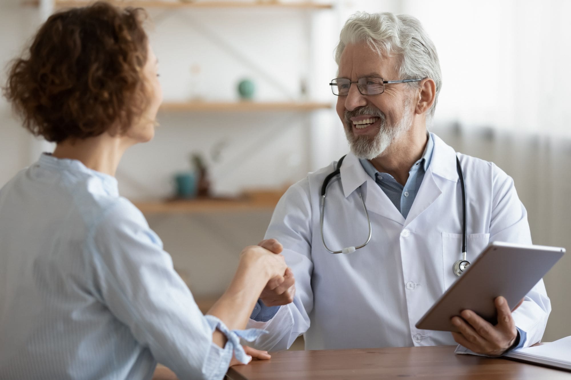 An older male doctor with gray hair and a beard smiles while shaking hands with a female patient. He is wearing a white coat, stethoscope, and holding a tablet. Both appear to be having a positive conversation.