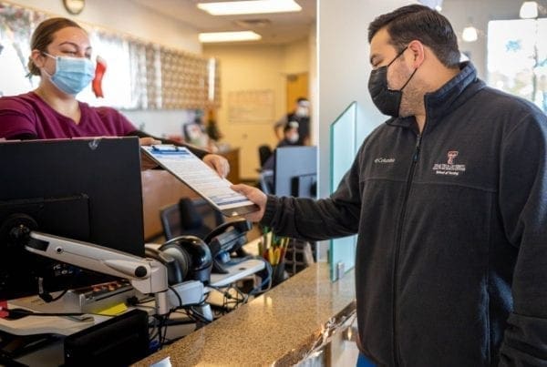 A man wearing a mask hands a clipboard to a masked receptionist at a front desk. The scene appears to be in an office or clinic, with a computer and office supplies on the counter.