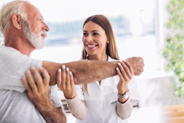 A smiling healthcare professional assists an older man by gently stretching his arm during a physical therapy session in a bright, well-lit room.