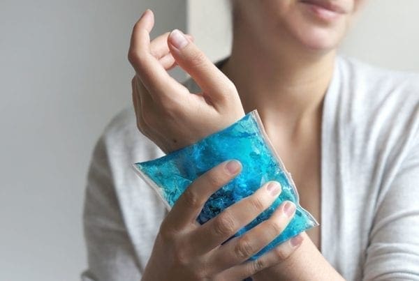 A person holds a blue gel ice pack against their wrist, likely to relieve pain or swelling. The person is wearing a light gray top and is visible from the shoulders down.