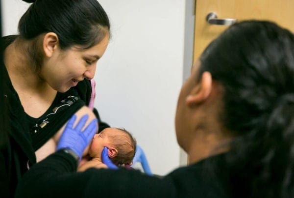 A woman smiles while a healthcare worker wearing blue gloves helps her breastfeed a newborn baby in a clinic room.