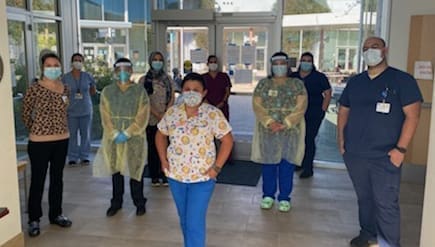 A group of healthcare workers wearing masks and scrubs, some in protective gowns, stand spaced apart in a hospital lobby with glass doors and daylight outside.
