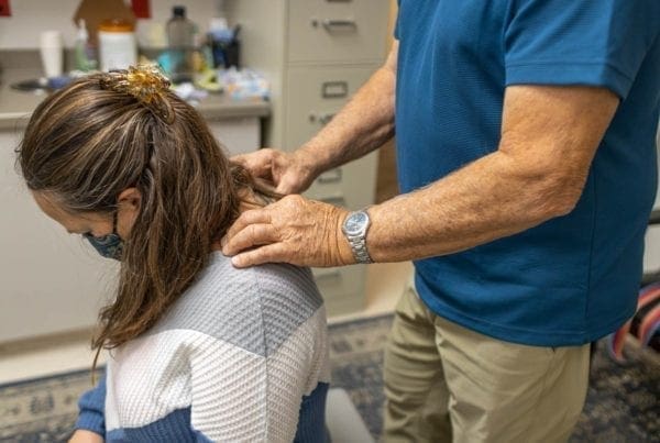 A person in a blue shirt gives a seated woman a neck and upper back massage in an office setting. The woman has a hair clip and is wearing a face mask. Office supplies and cabinets are visible in the background.