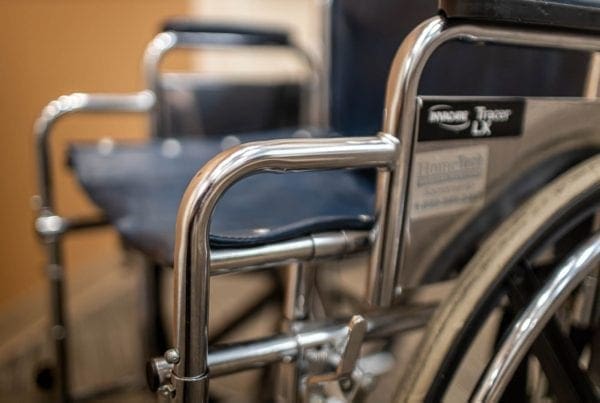 Close-up view of a wheelchairs metallic armrest and frame, with a second wheelchair blurred in the background. The image highlights the shiny metal and dark blue seat fabric.
