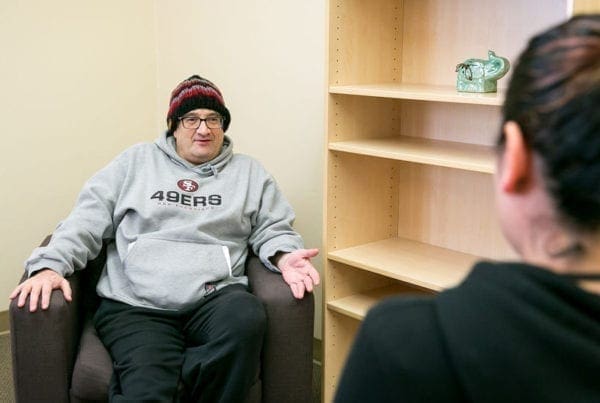 A person wearing a gray 49ers hoodie and a knit hat sits in an armchair, talking to another person whose back is to the camera, in an office with an empty wooden shelf.