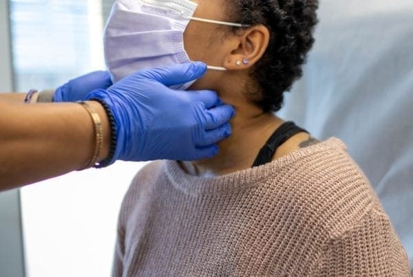A healthcare worker wearing blue gloves examines the neck of a woman sitting on an exam table. The woman wears a pink sweater and a face mask, and the setting appears to be a medical office.
