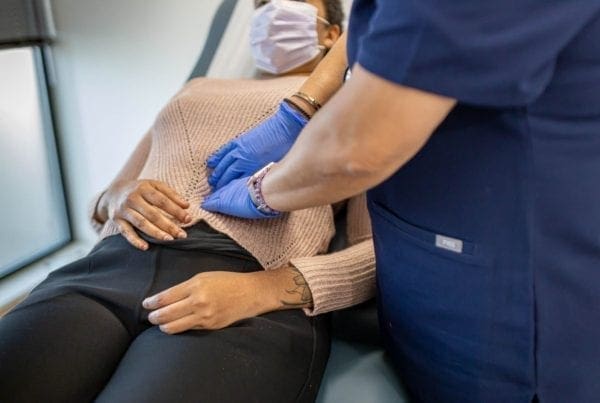 A person wearing a mask lies on an exam table while a healthcare worker in blue scrubs and gloves palpates their abdomen during a medical examination.