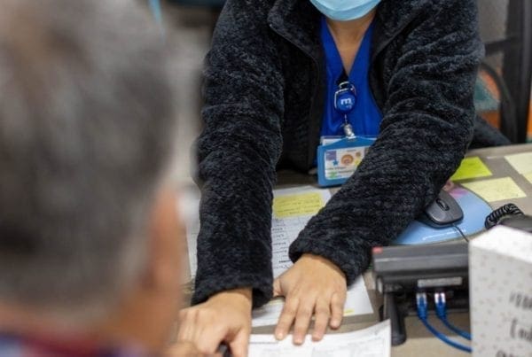 A woman wearing glasses and a face mask sits at a desk, pointing to a document in front of a man in a plaid shirt, likely explaining or reviewing paperwork with him in an office setting.