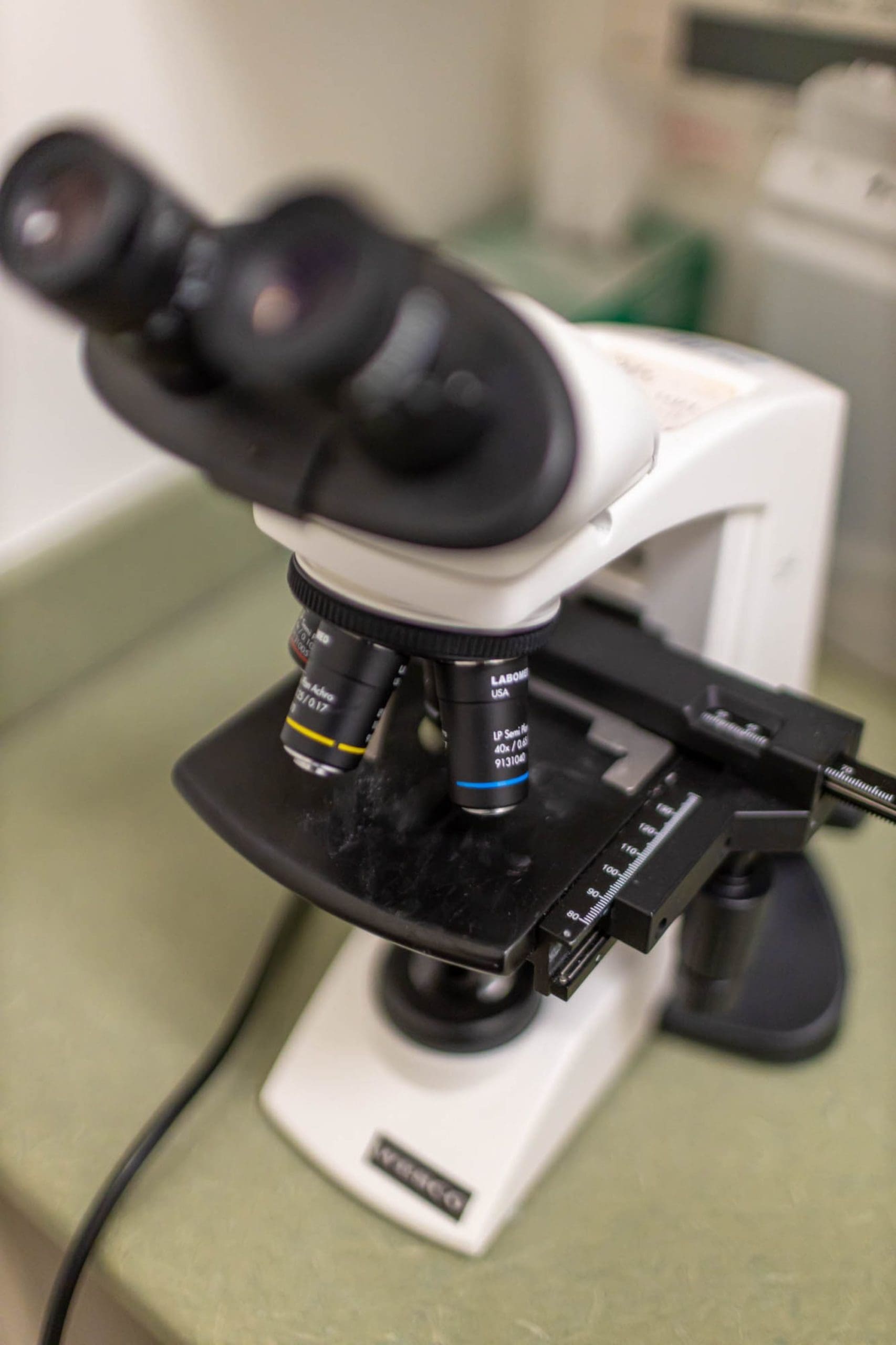 A close-up view of a laboratory microscope on a green countertop, showing the eyepieces, objective lenses, and stage in detail.