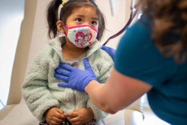 A young child wearing a colorful mask sits on a chair while a healthcare worker in gloves and a face shield gently places a hand on the childs chest during a medical checkup.