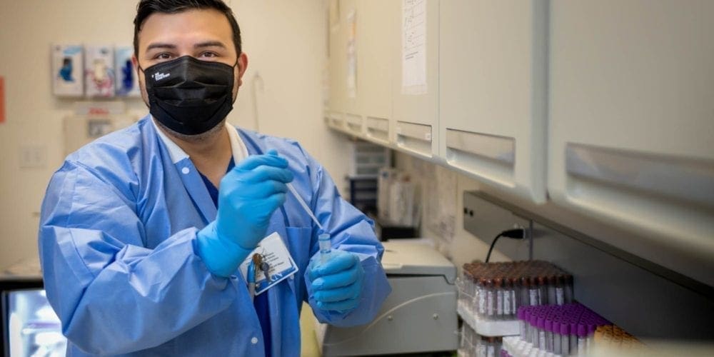 A lab technician wearing a black face mask, blue gloves, and a blue lab coat holds a pipette in a laboratory, with test tubes and equipment on the counter beside him.