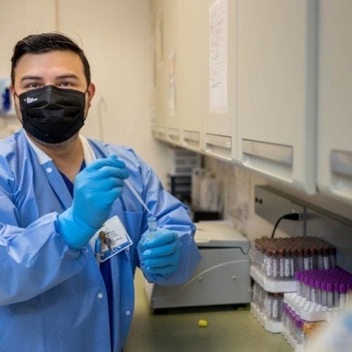 A lab technician wearing a black face mask, blue gloves, and a blue lab coat holds a pipette in a laboratory, with test tubes and equipment on the counter beside him.