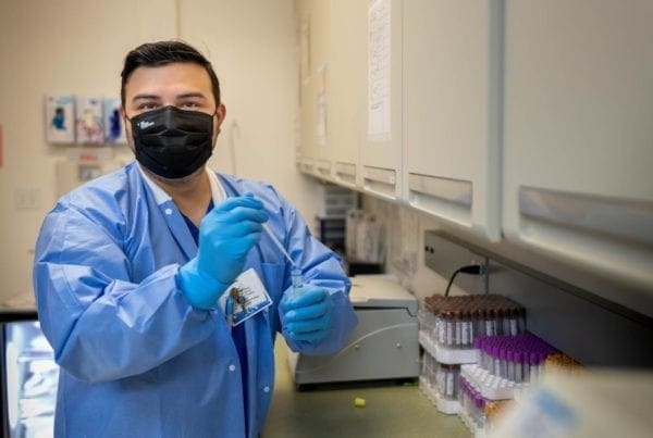 A lab technician wearing a black face mask, blue gloves, and a blue lab coat holds a pipette in a laboratory, with test tubes and equipment on the counter beside him.