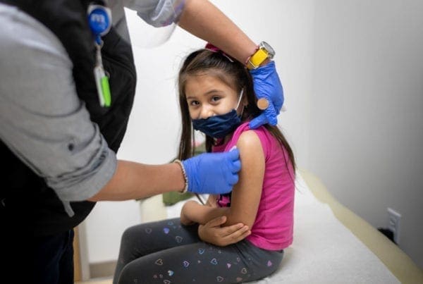 A young girl wearing a pink shirt and patterned leggings sits on an exam table while a healthcare worker in gloves prepares to give her a vaccine in her upper arm. The girl is smiling and wearing a partial face mask.