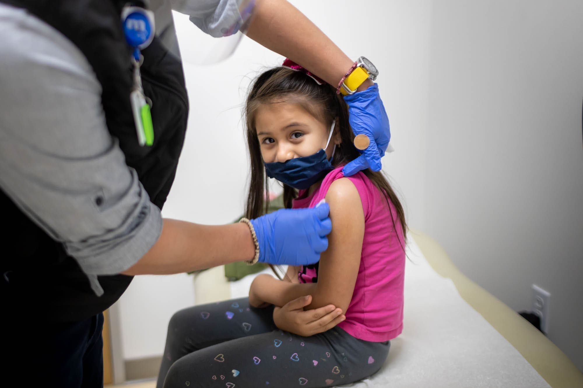 A young girl wearing a pink shirt and patterned leggings sits on an exam table while a healthcare worker in gloves prepares to give her a vaccine in her upper arm. The girl is smiling and wearing a partial face mask.