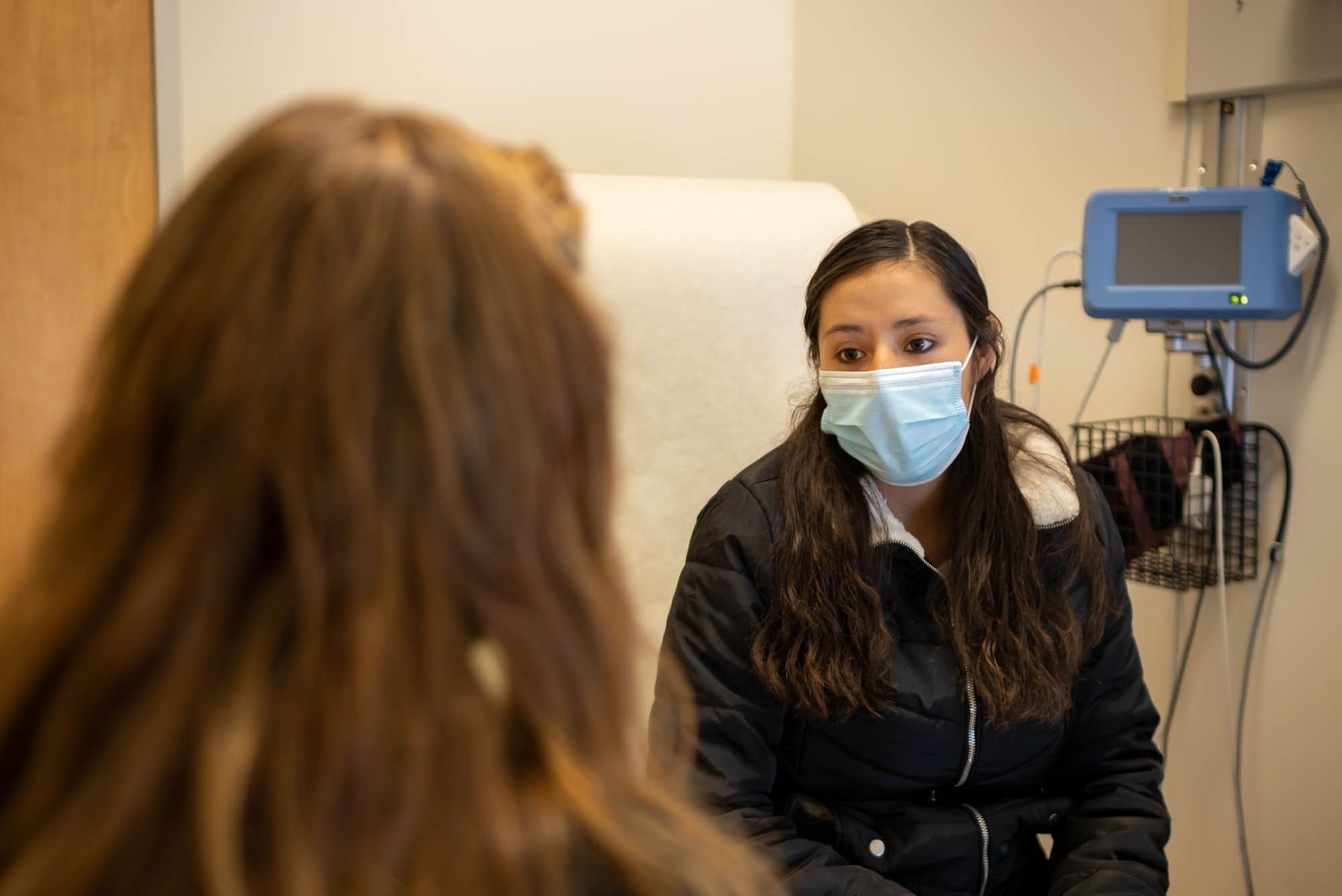 A young woman wearing a mask sits in a medical exam room, facing a healthcare professional whose back is to the camera. Medical equipment is visible on the wall.