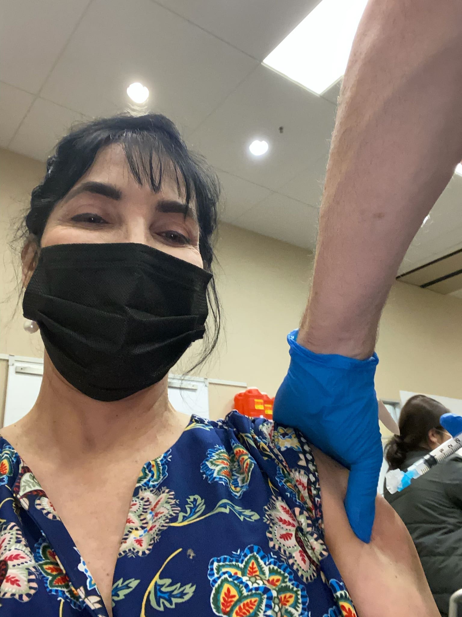 A woman in a black face mask receives a vaccine injection in her upper arm from a healthcare worker wearing blue gloves in a clinical setting.