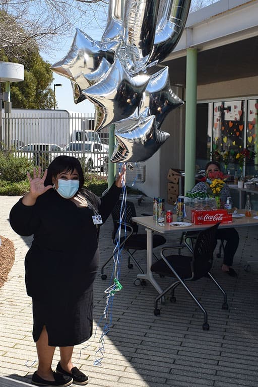 A woman in a black dress and face mask holds silver star-shaped balloons and waves, standing outside near a table with drinks and flowers. Another person sits at the table, and a building is in the background.