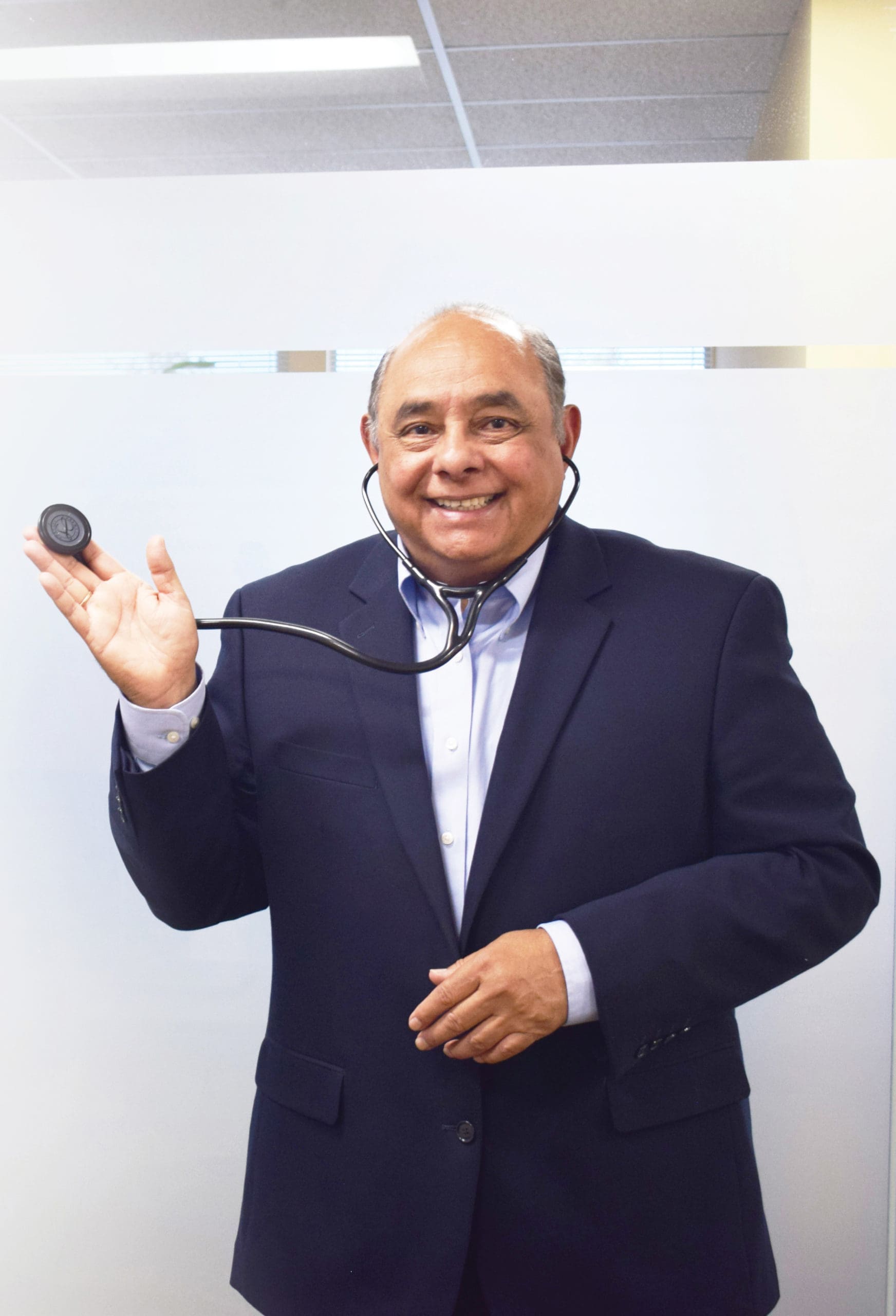 A smiling man in a navy blazer holds a stethoscope to his ear, standing indoors against a plain, light background.
