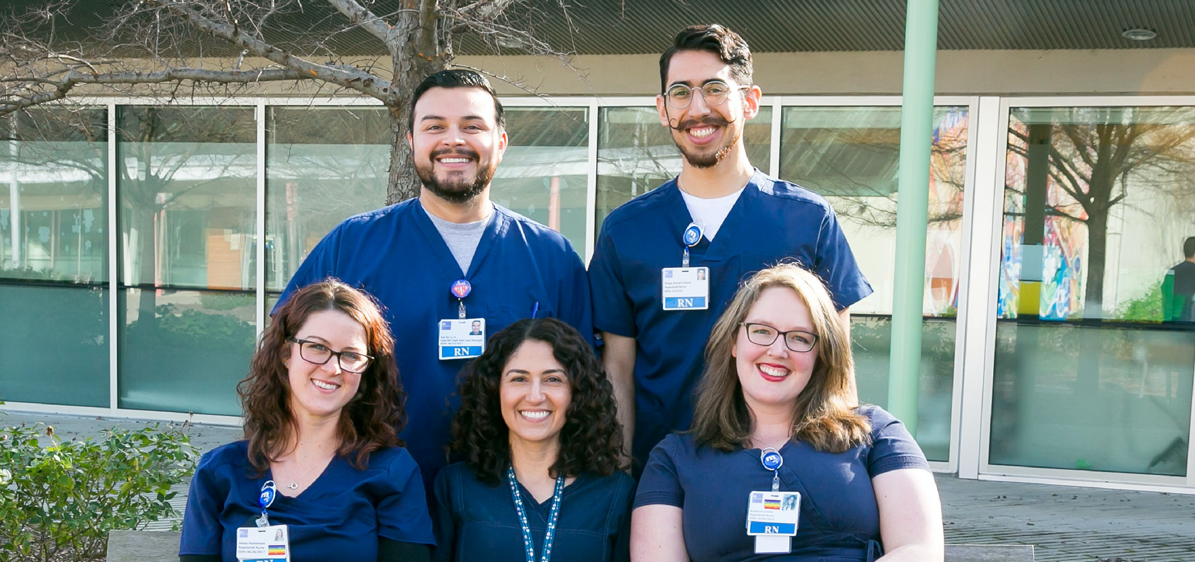 Five smiling healthcare professionals in blue scrubs and name badges pose together outdoors in front of a building with large windows and green plants.