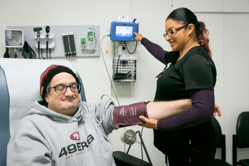 A healthcare worker in scrubs checks the blood pressure of a smiling man wearing glasses, a beanie, and a 49ers sweatshirt in a medical examination room.