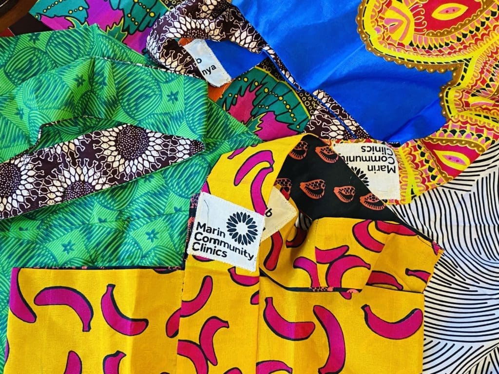 Colorful fabric masks with vibrant patterns, including green, yellow with pink shapes, blue, and black-and-white designs, are scattered overlapping each other. Some masks have Marin Community Clinics labels visible.