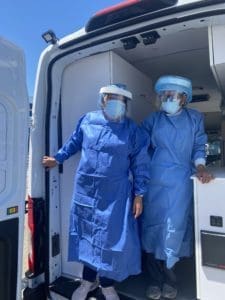 Two healthcare workers wearing blue gowns, face shields, masks, and gloves stand inside the open back of a medical van, preparing to step outside on a sunny day.
