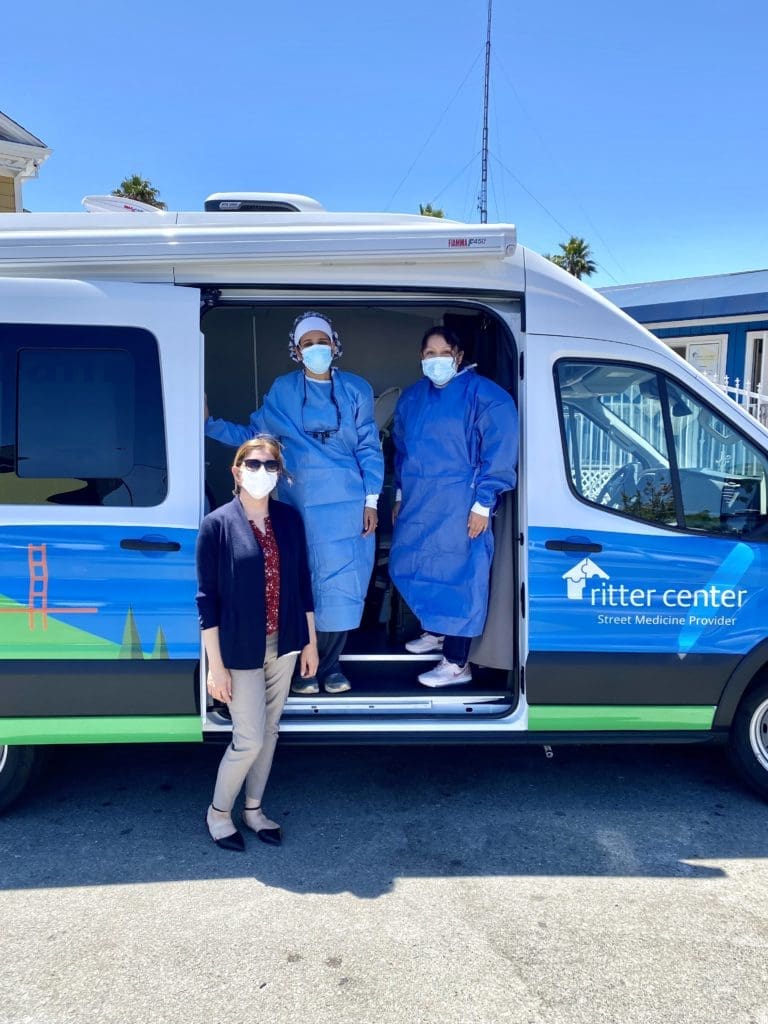 Three people wearing face masks and medical attire stand by a blue Ritter Center street medicine van; two are in blue gowns inside the van, and one stands outside, all appearing ready to provide healthcare services.