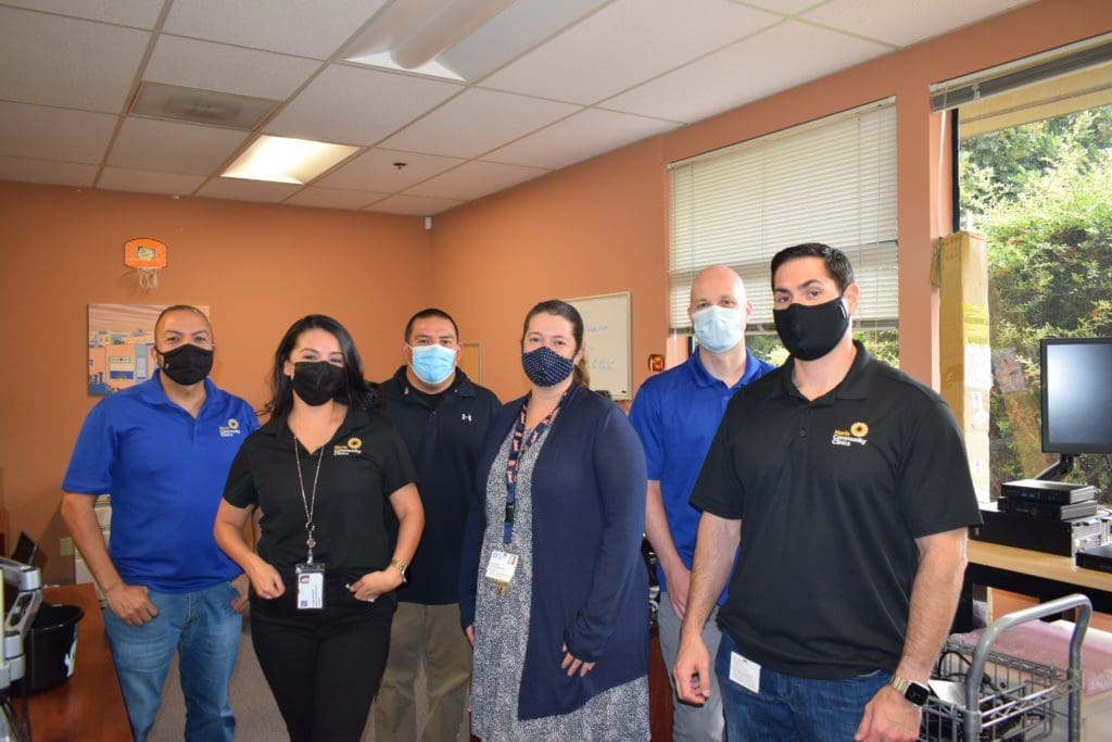 Six people, four men and two women, stand together in an office wearing face masks. They are dressed in business-casual attire, with ID badges visible, and are smiling for the camera under fluorescent lighting.