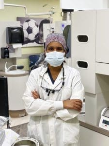 A dentist wearing a surgical mask, cap, and white coat stands with arms crossed in a dental office, with dental equipment and a soccer-themed wall decoration in the background.