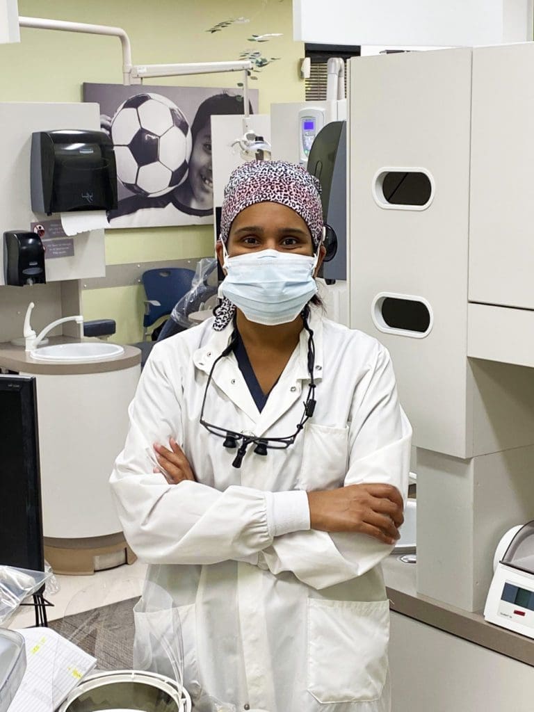 A dentist wearing a surgical mask, cap, and white coat stands with arms crossed in a dental office, with dental equipment and a soccer-themed wall decoration in the background.