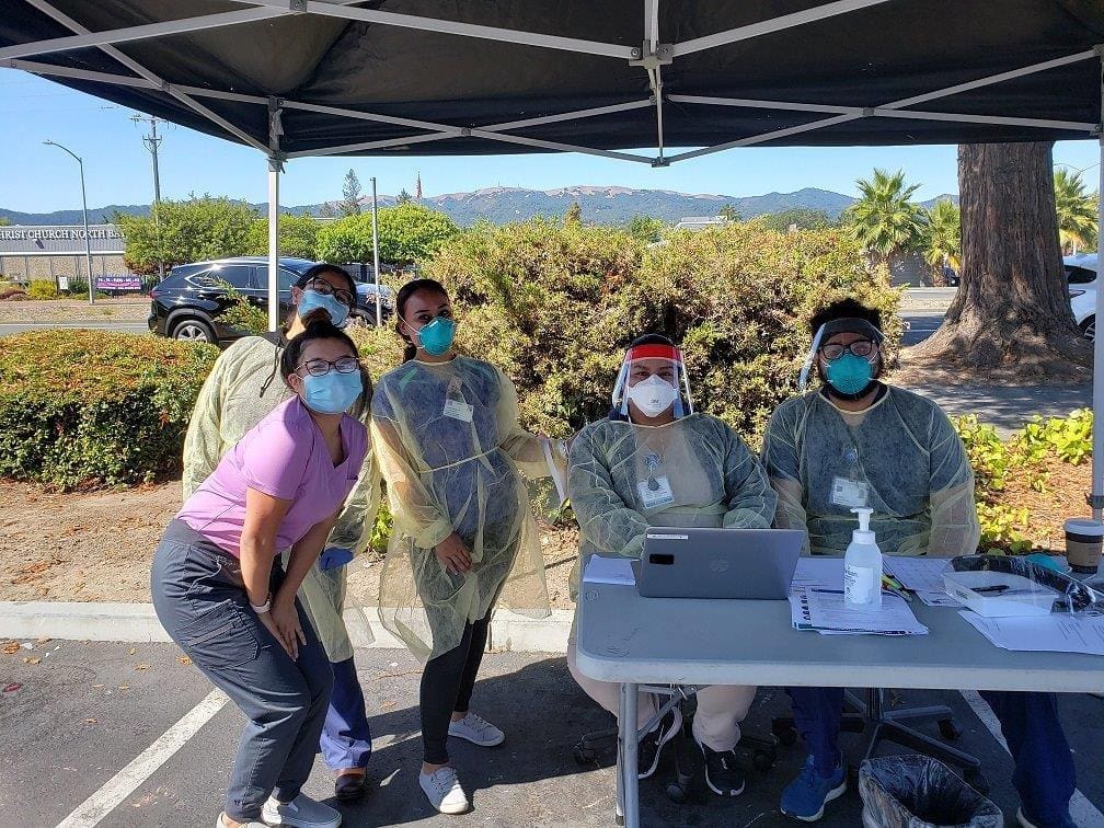 Five people in PPE, including masks and face shields, pose under a canopy outdoors. Two are seated at a table with a laptop and sanitizer, while three stand behind them. Trees and parked cars are visible in the background.