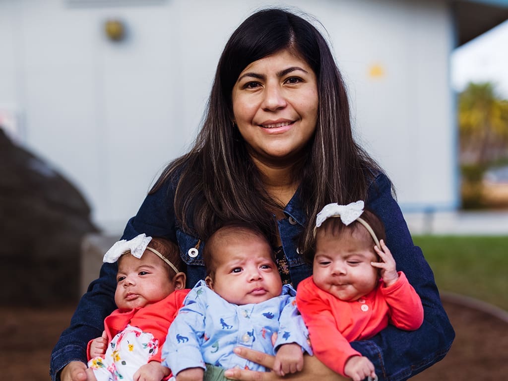 A woman with long dark hair smiles while holding three babies; two wear matching red outfits and white headbands, and the third wears a blue outfit with animal prints. They are outdoors with a blurred background.