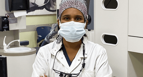 A healthcare worker wearing a mask, scrub cap, and white lab coat stands in a medical setting with arms crossed. Medical equipment and cabinets are visible in the background.