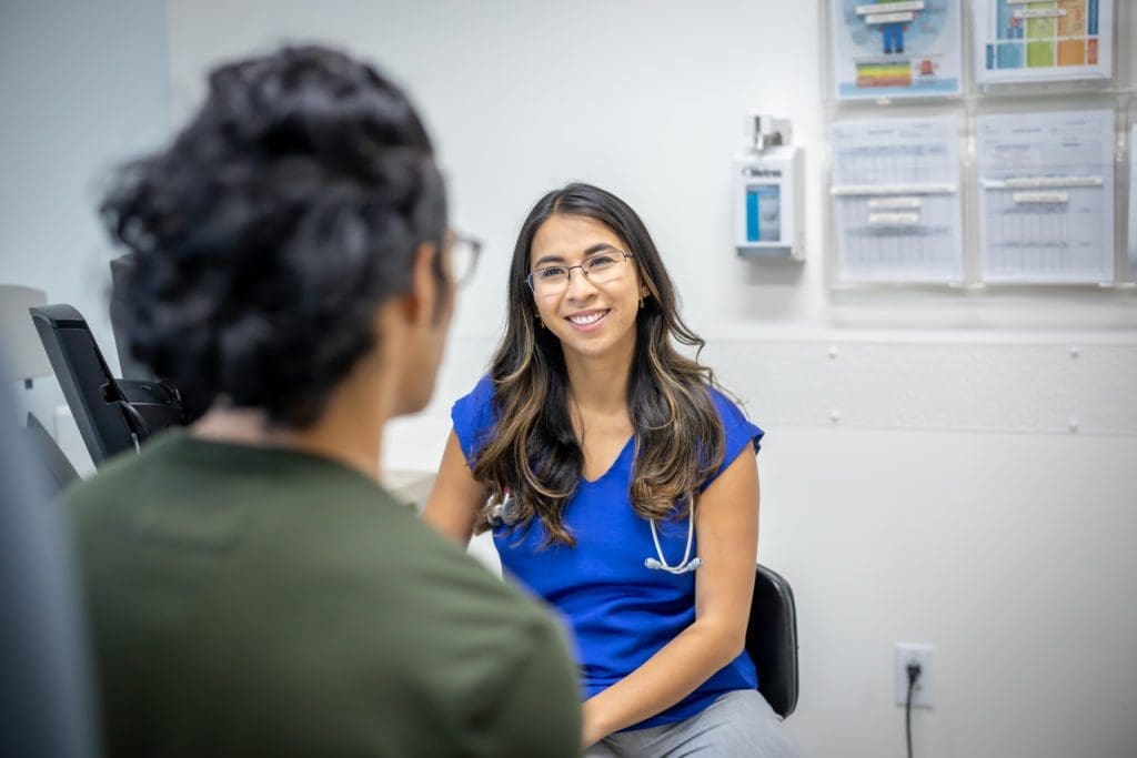 A female healthcare professional in blue scrubs and a stethoscope smiles while talking to a patient in a medical office. Charts and medical supplies are visible on the wall in the background.