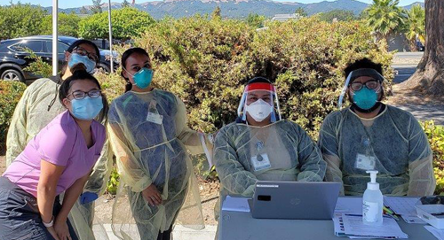 Five people wearing masks and protective gowns are outdoors at a table with a laptop and hand sanitizer. Some are seated, some standing, with bushes and mountains in the background. It appears to be a medical or testing site.