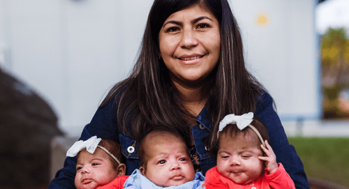 A woman with long dark hair smiles at the camera, holding three babies dressed in matching outfits and white bows on their heads. They are outdoors, with blurred greenery in the background.