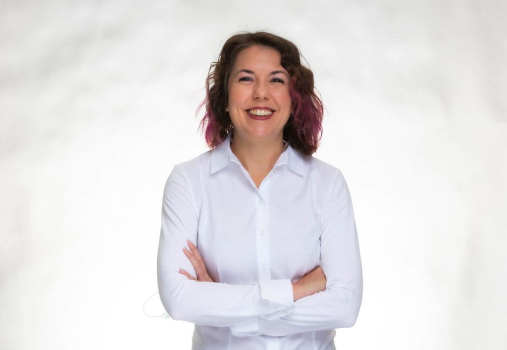 A woman with wavy brown hair and purple highlights smiles confidently with her arms crossed, wearing a white button-up shirt, standing against a plain white background.
