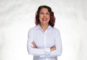 A woman with wavy brown hair and purple highlights smiles confidently with her arms crossed, wearing a white button-up shirt, standing against a plain white background.
