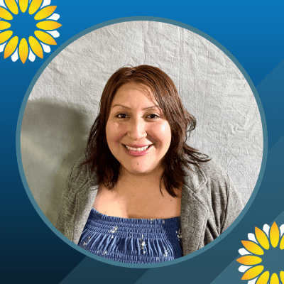 A woman with medium-length brown hair smiles at the camera. She is wearing a blue patterned top and a gray cardigan. The image has a blue border with yellow and white flower graphics in two corners.