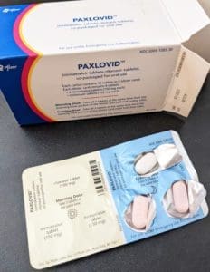 A box of Paxlovid and an opened blister pack showing pink oval tablets and white oval tablets, both labeled with dosages. The packaging includes usage instructions and emergency use authorization details.